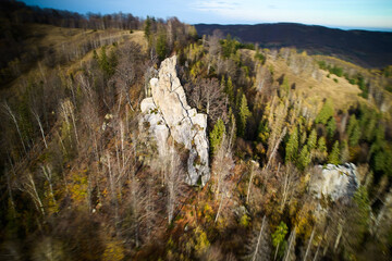 Dynamic aerial view of rocky spire rising sharply from forest floor. Surrounding landscape, with mix of bare trees and evergreen pines, creates striking contrast against the rugged rock formations.