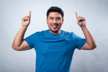 Fototapeta premium A smiling Asian man in a blue t-shirt raises his index finger as if making a point or presenting an idea. His confident and engaging expression conveys leadership, innovation, and inspiration