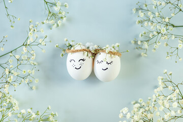 Two chicken eggs with hand drawn happy faces with wreaths and gypsophila branches on light blue...