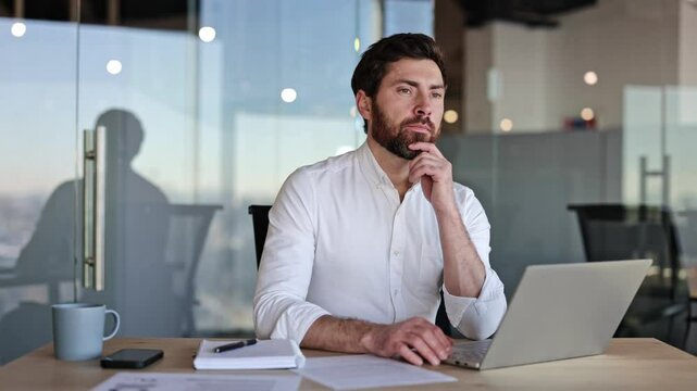 Focused businessman working on laptop in modern office. He thinks about project and types on keyboard.