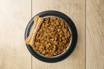 Dry pet food in feeding bowl and chew bone on wooden floor, top view