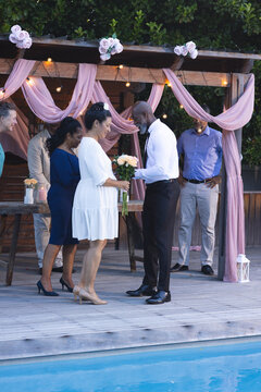 Smiling senior diverse couple exchanging flowers at in garden celebration with friends by poolside