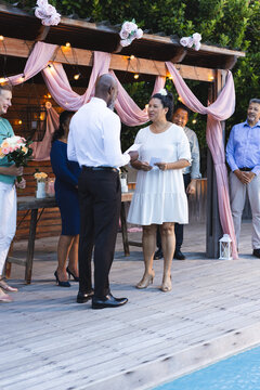 Senior diverse couple exchanging vows at in garden ceremony with friends smiling nearby