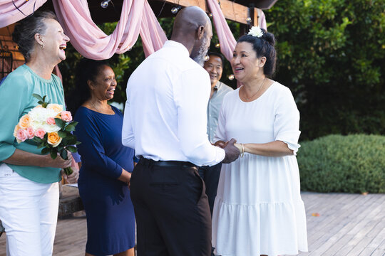 Celebrating engagement in garden, senior diverse couple holding hands with friends, smiling