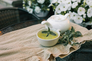 Cup of fresh herbal tea with mint leaves on a glass table. A white teapot and green herbs create a calming atmosphere. Hanging white flowers in the blurred background enhance the cozy outdoor setting