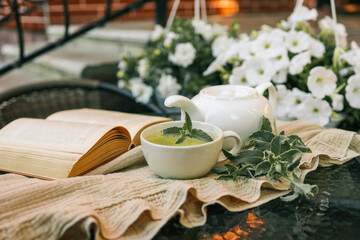 Cup of herbal tea with fresh mint on a cozy outdoor table. A white teapot, open book, and soft knit blanket create a relaxing atmosphere. White flowers in the background enhance the tranquil mood