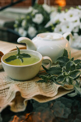 Cup of herbal tea with fresh mint on a cozy outdoor table. A white teapot, open book, and soft knit blanket create a relaxing atmosphere. White flowers in the background enhance the tranquil mood