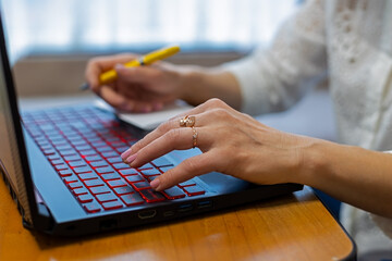 Close-up of woman's hands typing on a laptop keyboard, multitasking with a pen and notepad.