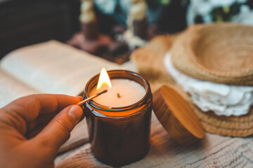 A hand lighting a wooden wick candle in an amber glass jar with a matchstick. Cozy setting with an...