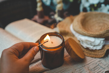 A hand lighting a wooden wick candle in an amber glass jar with a matchstick. Cozy setting with an open book, a straw hat, and soft fabric in warm, rustic lighting