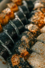Assorted sushi rolls in a takeout box with nori, sesame seeds, grilled cheese, and spicy topping. Japanese cuisine in warm sunlight with a close-up view of textures and details