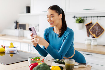 Smiling fit lady using cellphone while preparing healthy dinner and browsing food recipes for diet online, kitchen interior. Nutrition app concept