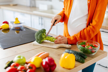 Pregnant woman cutting broccoli for healthy dinner, cooking at kitchen table filled with various vegetables, cropped, closeup