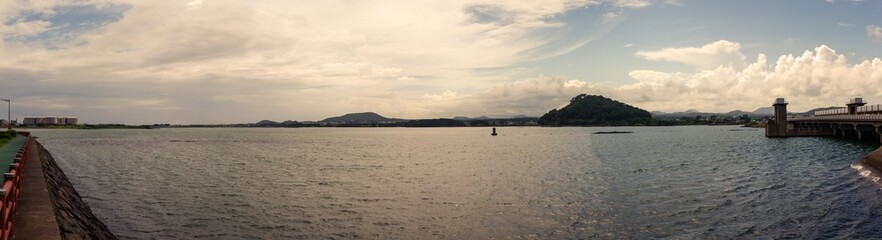 Naklejka premium Expansive Lakeside View with Calm Waters, Rolling Hills, and Cloud-Filled Sky Near Hallasan Mountain in Jeju