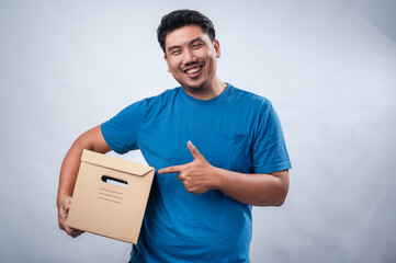 An Asian man in a blue t-shirt stands against a white background, smiling while holding a brown...