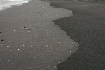 Black volcanic sand shoreline with scattered shells and smooth wet patterns shaped by receding ocean waves