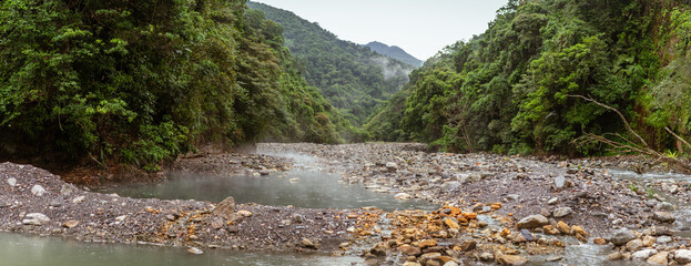 Thermal river meandering through lush green mountains with rising steam and rocky riverbanks in remote wilderness
