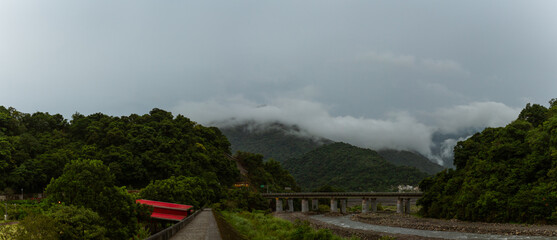 Mountain range covered in mist with lush greenery and river bridge under overcast sky in remote Taiwan region