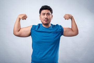 An Asian man in a blue sleeveless t-shirt flexes both arms, showing off his biceps in a strong, determined pose. His serious expression conveys power against a plain white background
