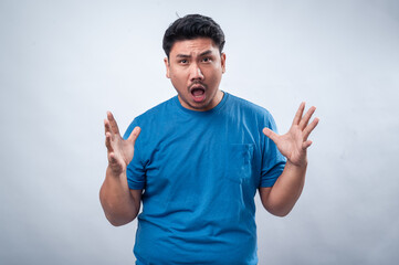 Fototapeta premium An Asian man in a blue t-shirt stands against a plain white background, raising both hands in a surprised and frustrated expression. His facial reaction conveys shock, disbelief, or confusion.