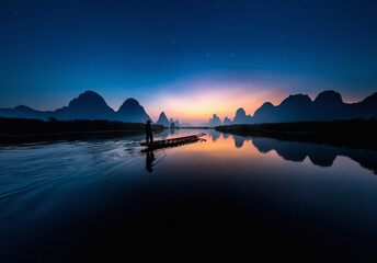 Serene dusk scene featuring a person on a bamboo raft in a calm river, with silhouetted mountains against a starry night sky.