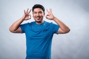 Naklejka premium A smiling Asian man in a blue t-shirt makes an OK hand gesture while looking confident and friendly. He stands against a clean white background, conveying positivity, approval, and encouragement.