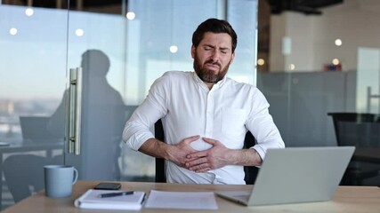 Bearded businessman in white shirt experiences painful stomach or abdomen ache while working on laptop in modern office.