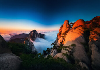Fototapeta premium Mountain peaks pierce through clouds at sunset. Golden light bathes rocky slopes as the sky blends blue and orange hues. A scenic, tranquil vista.
