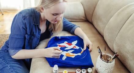 Female artist sitting on the couch and painting on fabric koi carp. Customization of clothes with...