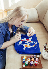 Female artist sitting on the couch and painting on fabric koi carp. Customization of clothes with...
