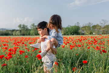 side view of piggyback ride of younger girl on elder child in a poppy field