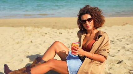 Sensual woman in denim summer clothes and bikini leaning on beach enjoying tropical fruit