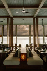 A stylish overwater restaurant with wooden interiors, ceiling fans, and glass railings, offering breathtaking ocean views and a serene dining experience at sunset in Maldives 