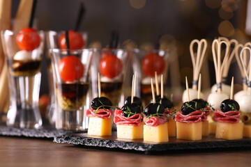 Many different tasty canapes on wooden table against blurred lights, closeup