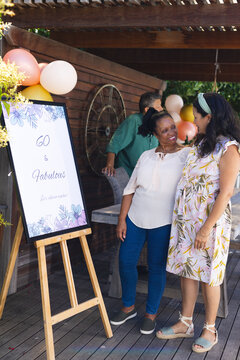Celebrating 60th birthday, two diverse female diverse senior friends smiling and enjoying outdoor pa