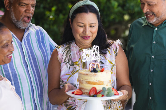 Celebrating 60th birthday, woman holding cake with diverse senior friends smiling outdoors - Powered by Adobe