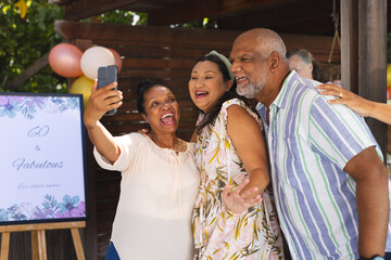 Diverse senior friends celebrating 60th birthday, taking joyful selfie at outdoor party