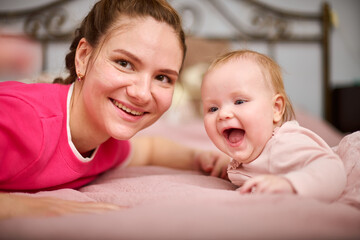 Young woman with brown hair and smiling baby girl on pink bed. Light-hearted interaction, creating joyful atmosphere. Warm lighting enhances bond