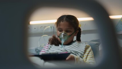 Little sick African American girl in pyjamas with oxygen mask on face sitting on hospitable bed and drawing while receiving treatment