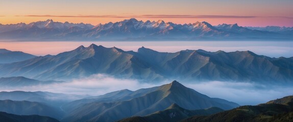 Panoramic view of misty mountain range at sunrise with snow-capped peaks and layered hills in soft light and fog in the valleys