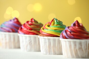 Delicious cupcakes with colorful cream on white table against golden background, closeup. Bokeh effect