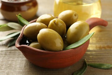 Tasty green olives and leaves in bowl on wooden table, closeup