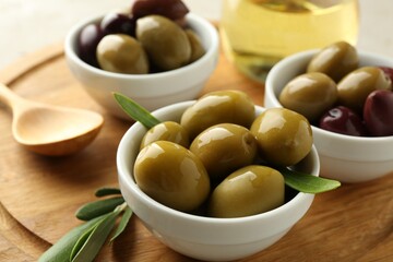 Delicious marinated olives in bowls, spoon, oil and green leaves on table, closeup