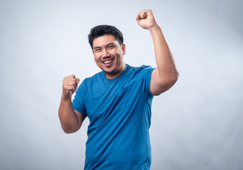 Fototapeta premium An Asian man in a blue t-shirt clenches his fists and shows an intense expression of excitement or triumph against a white background. His face conveys determination and strong emotions