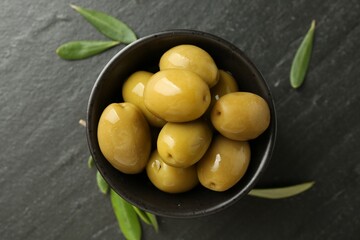 Delicious marinated olives in bowl and green leaves on black table, flat lay