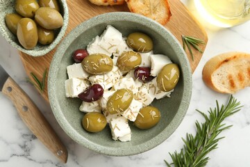 Marinated olives with feta cheese, bread pieces, oil and rosemary on white marble table, flat lay