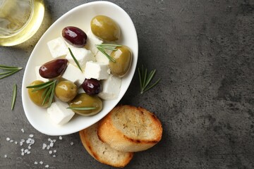 Marinated olives with feta cheese, bread pieces, oil, salt and rosemary on grey table, flat lay. Space for text
