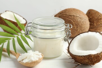 Organic coconut cooking oil in jar, spoon, fresh fruits and green branch on white wooden table, closeup