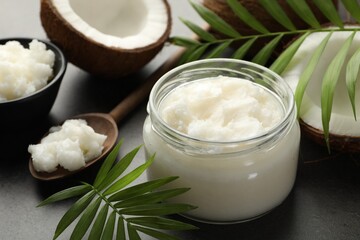 Organic coconut cooking oil in jar, spoon, fresh fruits and green branches on grey table, closeup