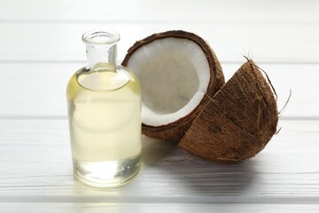Organic coconut cooking oil in glass bottle and halves of fruit on white wooden table, closeup